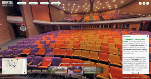 A screenshot of Bristol Community College's Margaret L. Jackson Arts Center on their 360° Tour. A large, brick-walled, brightly-lit theater facing orange, purple, and yellow two-leveled seating.