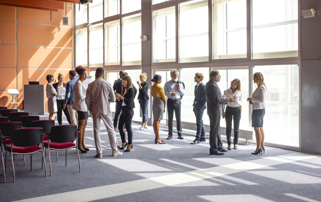 A group of diverse business executives are engaging in conversations at a business conference. They are standing in a spacious, sunlit room with large windows and modern decor, fostering a professional networking environment.